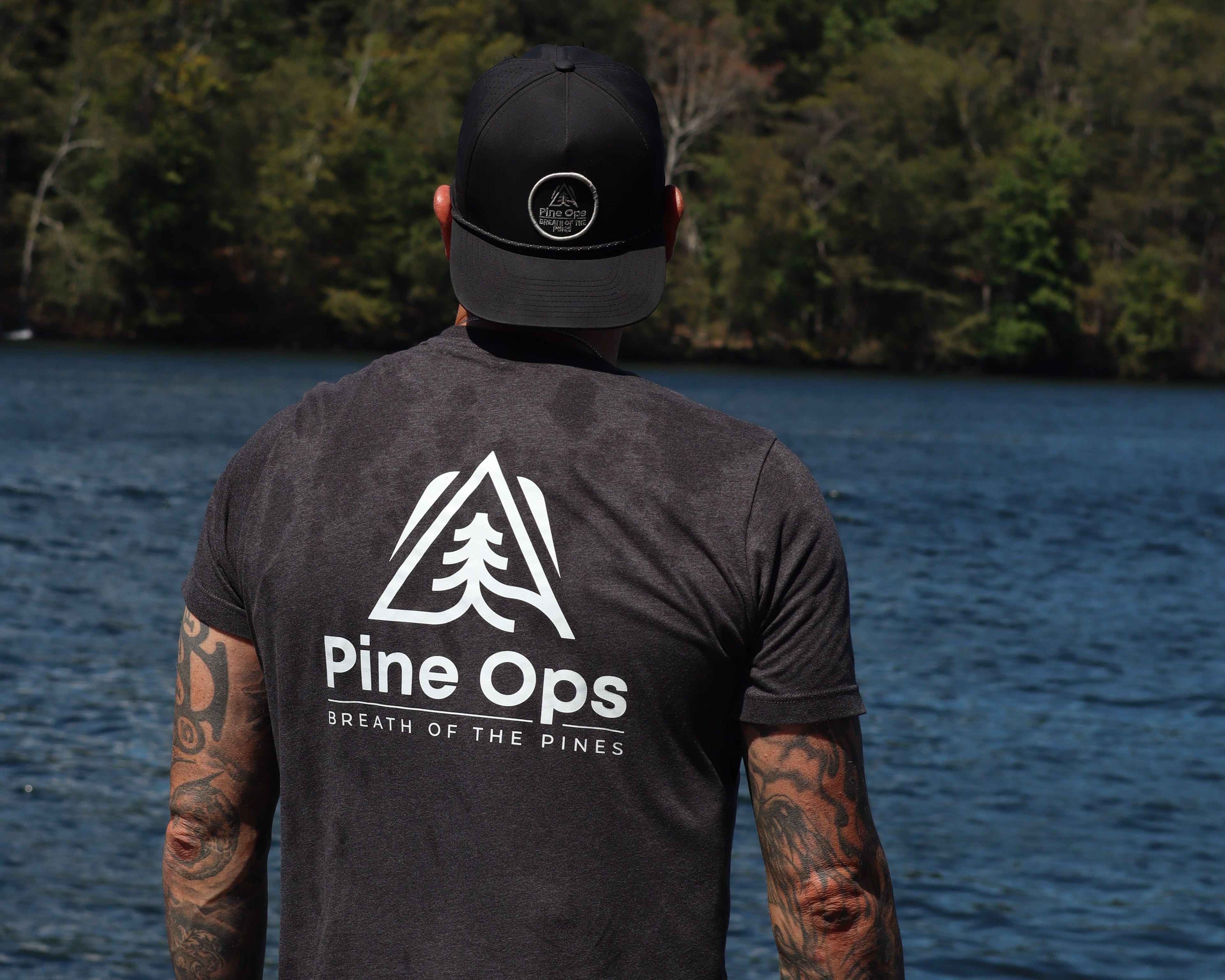 Person wearing a black Pine Ops shirt and cap by a lake with trees in the background
