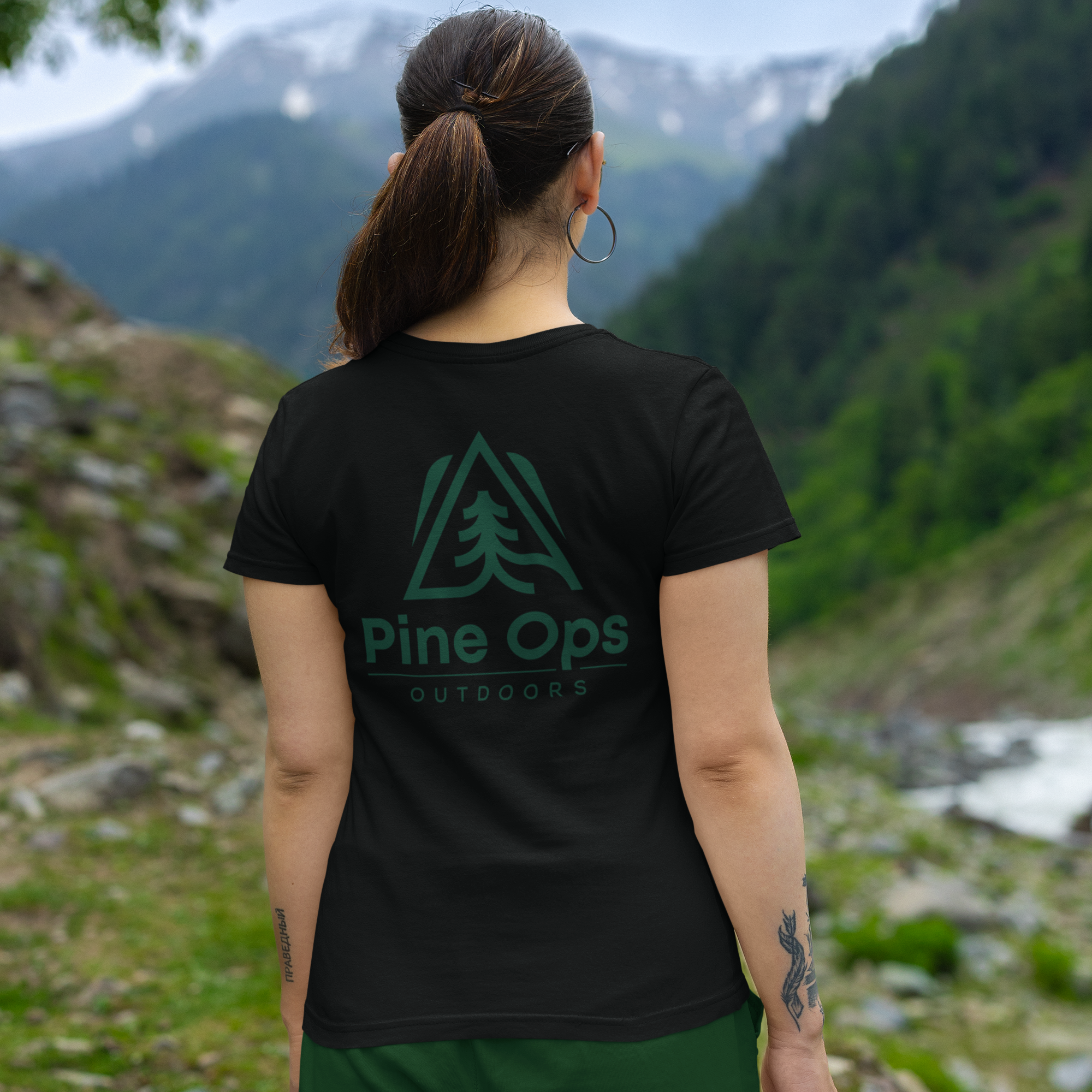 Person wearing a black t-shirt with 'Pine Ops Outdoors' logo in a mountainous landscape